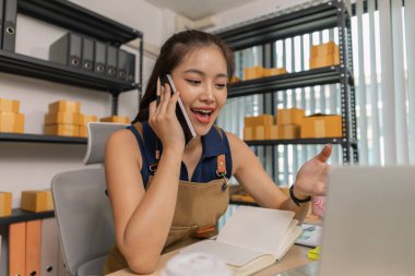 Genç bayan girişimci telefonda konuşurken gülümsüyor ve evde dizüstü bilgisayar kullanıyor. Bir internet mağazası işletiyor, siparişleri kontrol ediyor ve e-ticaret işi için müşterilerle iletişim kuruyor..