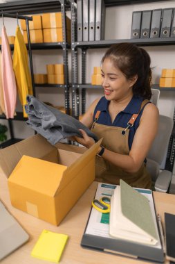 Gülümseyen genç bayan girişimci giysilerini bir paket kutusuna koyup, mutlu bir şekilde sipariş hazırlıyor, organize edilmiş çalışma alanından başarılı online moda mağazasını yönetiyor..
