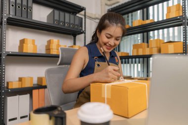 Genç bayan girişimci bir paket etiketinin üzerine bir nakliye adresi yazarak ev ofisine teslimat için bir paket hazırlıyor. Online bir iş yürütüyor ve müşterilere sevkiyat düzenliyor..