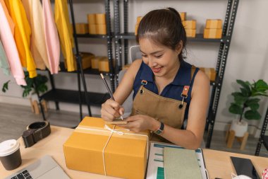 Genç bayan girişimci bir paket etiketinin üzerine bir nakliye adresi yazarak ev ofisine teslimat için bir paket hazırlıyor. Online bir iş yürütüyor ve müşterilere sevkiyat düzenliyor..