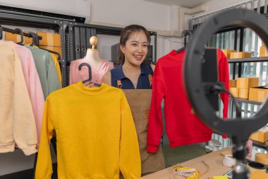 Genç bayan girişimci, ev ofisinden kıyafet satmak için yaşarken gülümsüyor. Moda ürünleri sunar, izleyicilerle iletişim kurar ve canlı seans sırasında online mağazasını yönetir..