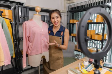 Genç bayan girişimci, ev ofisinden kıyafet satmak için yaşarken gülümsüyor. Moda ürünleri sunar, izleyicilerle iletişim kurar ve canlı seans sırasında online mağazasını yönetir..