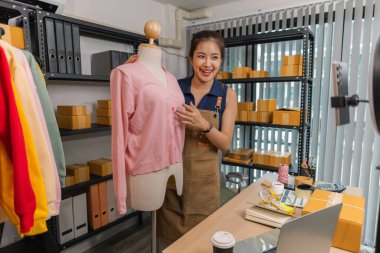 Genç bayan girişimci, ev ofisinden kıyafet satmak için yaşarken gülümsüyor. Moda ürünleri sunar, izleyicilerle iletişim kurar ve canlı seans sırasında online mağazasını yönetir..