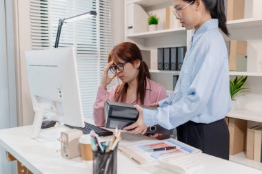 Bir ofiste çalışan iki bayan meslektaşı, biri eli alnında stresli görünürken diğeri de yoğun bir departmanda takım çalışmasını ve iş baskısını sembolize eden bir klasör sunuyor..