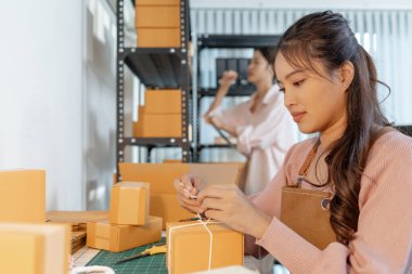 Küçük bir işletme ofisinde birlikte çalışan iki genç kadın, karton kutular hazırlıyor, envanter kontrolü yapıyor ve yeni açılan çevrimiçi mağaza operasyonlarının bir parçası olarak ambalaj işlerini yürütüyorlar..