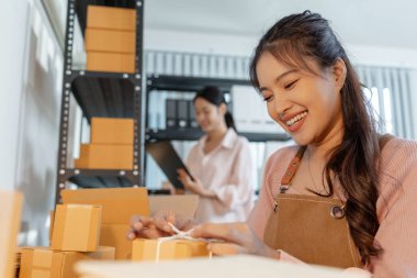 Küçük bir işletme ofisinde birlikte çalışan iki genç kadın, karton kutular hazırlıyor, envanter kontrolü yapıyor ve yeni açılan çevrimiçi mağaza operasyonlarının bir parçası olarak ambalaj işlerini yürütüyorlar..