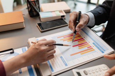 Modern bir ofis masasında toplanan iş adamları, planlar ve bütçe raporlarını gözden geçirirken, aynı zamanda yaklaşan iş kararları için temel mali içgörüler ve stratejik planlar üzerinde de tartıştılar..