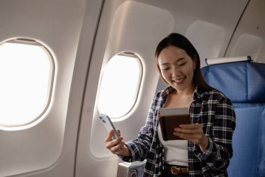 Pasaportu ve akıllı telefonu olan genç bir Asyalı kadın, seyahat belgelerini incelerken gülümsüyor ve ekonomi sınıfı kabininde uçuş deneyiminin tadını çıkarıyor..