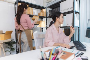 İki genç kadın, kargo kutuları ve belgelerle çevrili küçük bir iş ofisinde birlikte çalışıyorlar. Siparişleri tartışıyorlar, paketleri hazırlıyorlar ve envanteri bilgisayarda yönetiyorlar..