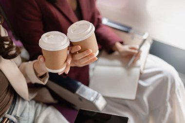 İki kadın uçakta birlikte oturur, gülümser ve pencerenin kenarında kahve fincanları tutarlar, arkadaşça etkileşim kurarlar, seyahat anlarını paylaşırlar, ve uçuş sırasında rahat bir atmosferi paylaşırlar..