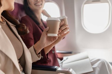 İki kadın uçakta birlikte oturur, gülümser ve pencerenin kenarında kahve fincanları tutarlar, arkadaşça etkileşim kurarlar, seyahat anlarını paylaşırlar, ve uçuş sırasında rahat bir atmosferi paylaşırlar..