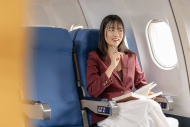 Genç bir kadın uçak penceresinin kenarında oturur, sakince okur ve not alır. Odaklanmış kişisel zamanı, sessiz üretkenliği ve rahat bir hava yolculuğu deneyimi sırasında huzurlu bir anı temsil eder..