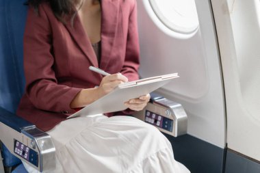 Genç bir kadın uçak penceresinin kenarına oturmuş, dikkatle bir panoya yazı yazıyor, sessiz üretkenliği, kişisel konsantrasyonu ve hava yolculuğu sırasında sakin çalışma anını temsil ediyor..