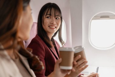 İki kadın uçakta birlikte oturur, gülümser ve pencerenin kenarında kahve fincanları tutarlar, arkadaşça etkileşim kurarlar, seyahat anlarını paylaşırlar, ve uçuş sırasında rahat bir atmosferi paylaşırlar..