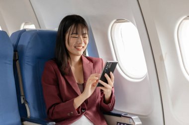 Genç bir kadın elinde akıllı telefon ve bir fincan kahveyle uçağın penceresinde oturur, sakin bir anın, modern seyahat rahatlığının ve rahat bir uçuş süresinin tadını çıkarır..