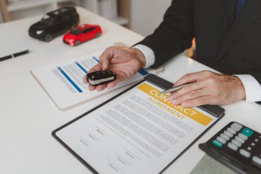 Resmi takım elbiseli bir işadamı, araba sigortası detaylarını, küçük kırmızı bir araba modelini elinde tutarak ve sözleşme belgelerine işaret ederek, anlaşmayı, korumayı ve mali planlamayı sembolize ederek açıklıyor.