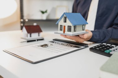 Bir işadamı ev modelleri, anahtarlar ve sözleşme belgeleri, emlak yatırımları ve ipotek anlaşmalarının altını çizerek ev sahipliği, finans ve emlak başarısını sembolize ediyor.