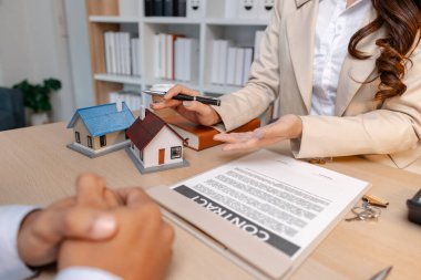 Bir emlakçı ve müvekkil ofis masasında emlak satış detaylarını tartışıyor, minyatür ev modelleri ve masanın üzerindeki parayla ev satın alma sözleşmelerini gözden geçiriyor ve imzalıyor..