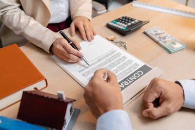 Bir emlakçı ve bir müşteri, emlak satın alma toplantısı sırasında bir konut sözleşmesi belgesini tartışıyor ve ona ev sahipliği hakkında anlaşma detayları ve mali müzakereleri gösteriyorlar.