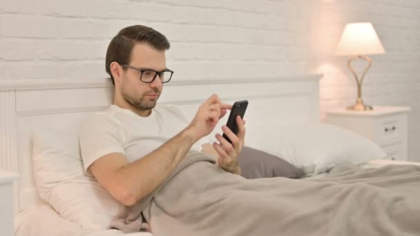 Hombre joven usando Smartphone en la cama — Vídeos de Stock