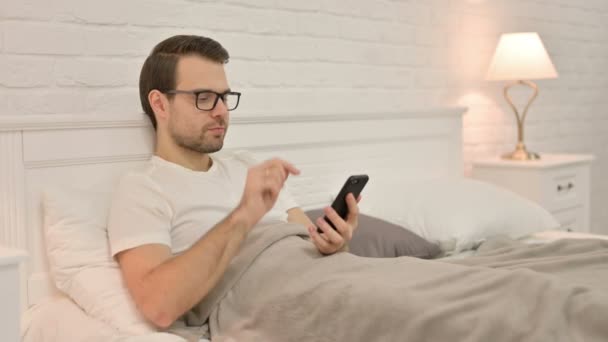 Succes, Jonge man viert feest op smartphone in bed — Stockvideo