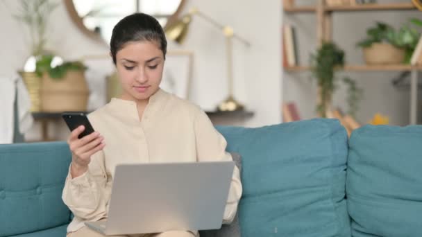 Młoda Hinduska kobieta pracuje na smartfonie i laptopie na kanapie — Wideo stockowe