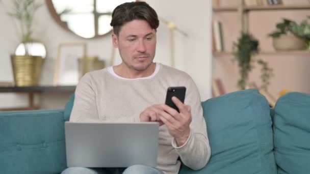 Среднего возраста человек со смартфоном работает на ноутбуке, на дому — стоковое видео