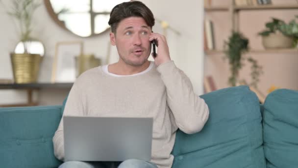 Hombre de mediana edad con el ordenador portátil hablando en el teléfono inteligente, en casa — Vídeos de Stock