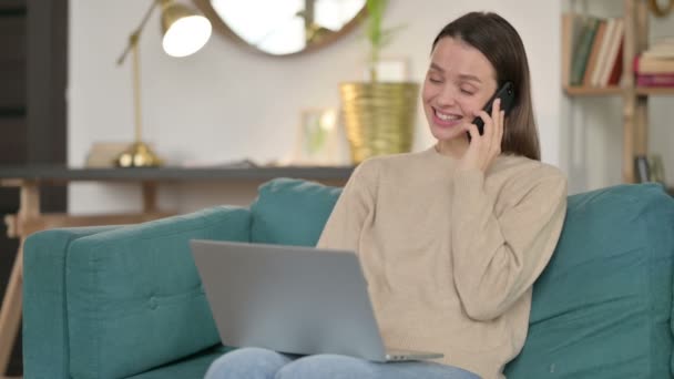 Jonge vrouw met laptop praten op smartphone op sofa — Stockvideo