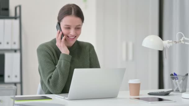 Mujer joven con el ordenador portátil hablando en el teléfono inteligente — Vídeo de stock