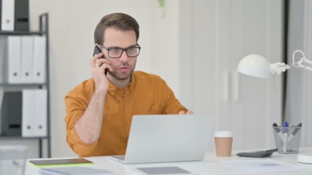 Hombre joven con el ordenador portátil que habla en el teléfono inteligente — Vídeos de Stock