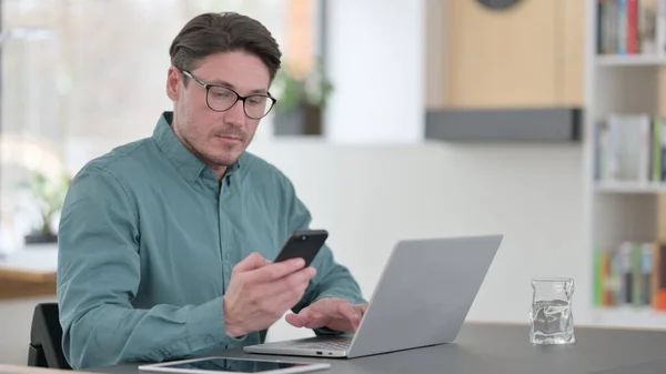 Hombre de mediana edad utilizando Smartphone mientras trabaja en el ordenador portátil —  Fotos de Stock
