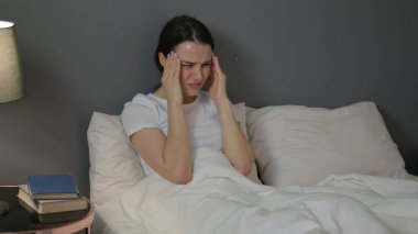 Başağrısı olan genç bir kadın. Yatakta oturuyor. 