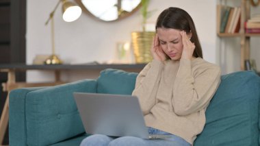 Dizüstü bilgisayarlı genç kadın koltukta baş ağrısı çekiyor. 