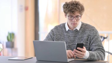 Genç adam iş yerinde akıllı telefon ve dizüstü bilgisayar kullanıyor. 