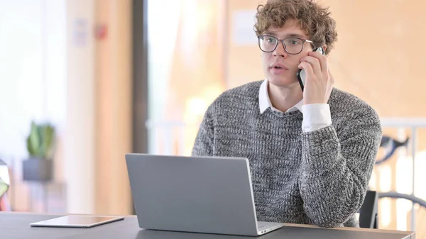 Hombre joven con el ordenador portátil hablando en el teléfono inteligente en el trabajo —  Fotos de Stock