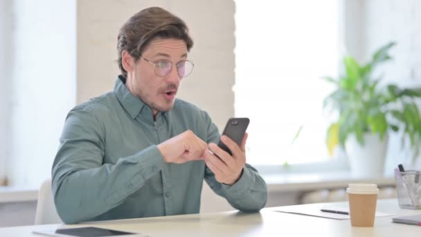 Чоловік середнього віку реагує на втрати на смартфон в офісі — стокове відео