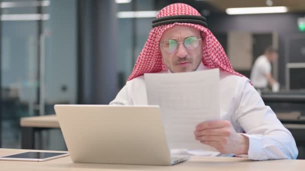 Hombre árabe de mediana edad con computadora portátil reaccionando a documentos de pérdida — Vídeo de stock