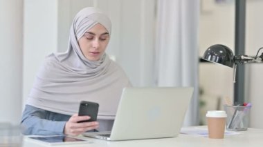 Ofiste akıllı telefon kullanan dizüstü bilgisayarlı genç bir Arap kadın.