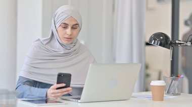 Ofiste akıllı telefon kullanan dizüstü bilgisayarlı genç bir Arap kadın.