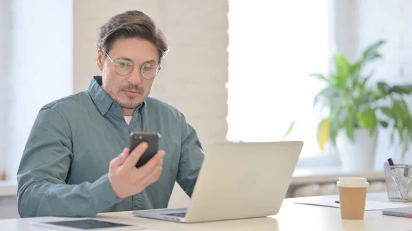 Hombre de mediana edad con ordenador portátil usando Smartphone en la oficina — Foto de Stock