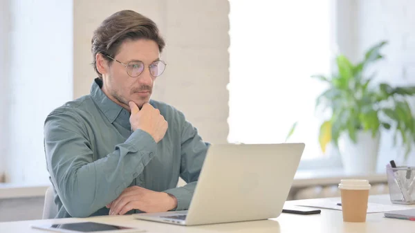 Hombre de mediana edad Pensando mientras usa el ordenador portátil en la oficina —  Fotos de Stock