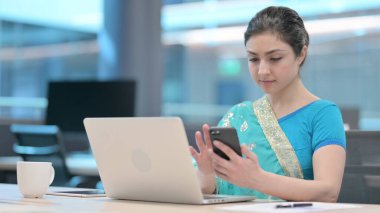 İş yerinde akıllı telefon kullanan dizüstü bilgisayarlı Hintli kadın.