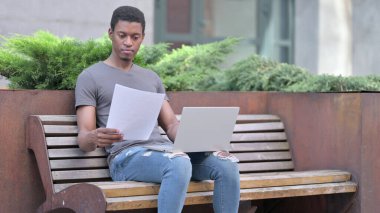 Genç Afrikalı Adam Bench 'te kağıt ve dizüstü bilgisayar üzerinde çalışıyor. 
