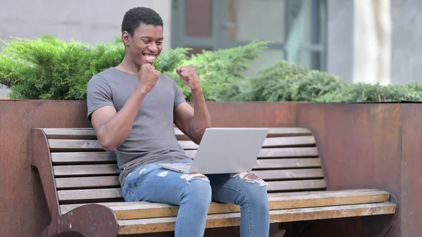 Spännande ung afrikansk man firar framgång på laptop på bänk — Stockfoto