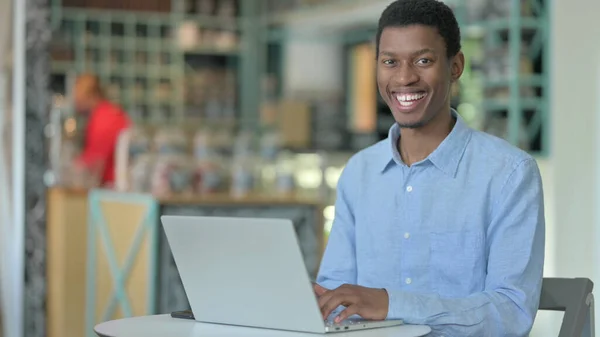 Ung afrikansk man med bärbar dator ler mot kameran i Café — Stockfoto