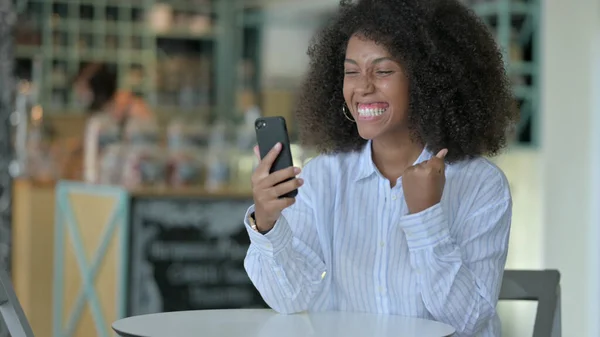 उत्तेजित युवा अफ्रीकी महिला कैफे में स्मार्टफ़ोन पर सफलता मना रही — स्टॉक फ़ोटो, इमेज
