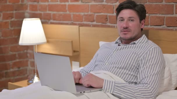 Hombre de mediana edad con ordenador portátil sonriendo a la cámara en la cama — Vídeos de Stock