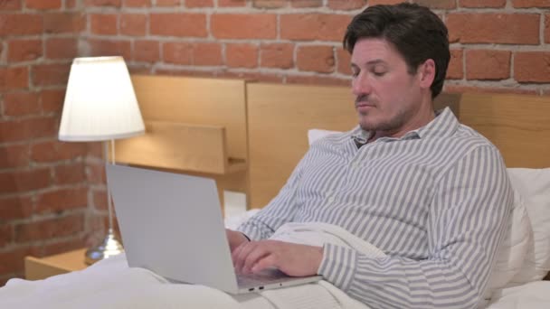 Mann mittleren Alters zeigt mit Laptop auf die Kamera im Bett — Stockvideo