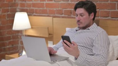 Orta yaşlı adam yatakta uyurken akıllı telefon kullanıyor.